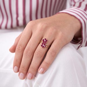 Puede incluir: Un anillo de oro con dos motivos en forma de flor, cada uno engastado con cinco piedras preciosas de color rosa intenso en forma de p&eacute;talo y una peque&ntilde;a piedra redonda transparente en el centro. El aro es fino y pulido.