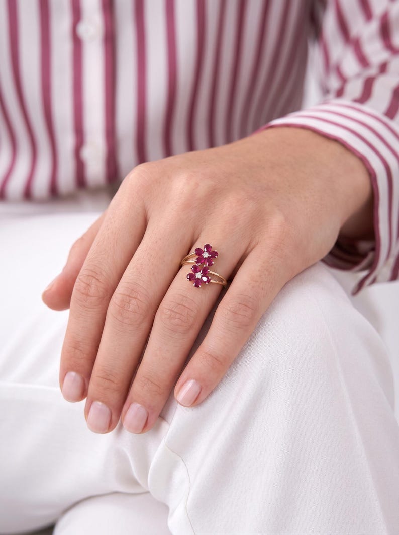 Puede incluir: Un anillo de oro con dos motivos en forma de flor, cada uno engastado con cinco piedras preciosas de color rosa intenso en forma de p&eacute;talo y una peque&ntilde;a piedra redonda transparente en el centro. El aro es fino y pulido.