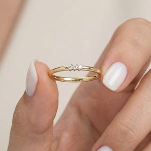 Könnte beinhalten: Ein zarter goldener Ring mit drei kleinen, klaren Edelsteinen in einer Reihe. Der Ring hat ein dünnes Band und das Wort "FIOR" ist innen eingraviert. Der Ring wird von einer Person gehalten.