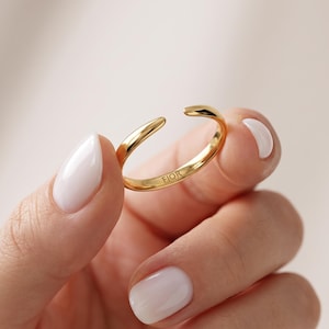 Könnte beinhalten: Ein goldfarbener offener Ring mit einer glatten, polierten Oberfläche. Die Innenseite des Rings ist deutlich mit dem Text FIOR graviert. Das Design besteht aus einem einfachen, durchgehenden Metallband mit zwei spitz zulaufenden Enden.