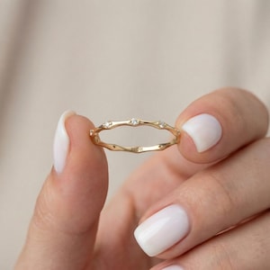 Peut inclure: Une bague en or délicate avec un design ondulé unique. La bague présente de petites pierres précieuses scintillantes espacées uniformément le long de l'anneau. Le mot "FLOR" est gravé sur la bague. La bague est tenue entre deux doigts.