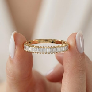 May include: A gold ring featuring a band of rectangular-cut diamonds. The ring is held between two fingers. The word "FIOR" is engraved on the ring. The ring is a piece of jewellery.