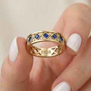 May include: A gold ring with a repeating pattern of square blue gemstones and small clear stones. The word "FIOR" is engraved on the ring's band. The ring is held between two hands with white nail polish.