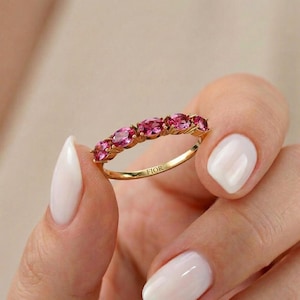 May include: A gold ring with a line of oval pink gemstones. The ring is held in a hand with white nail polish. The word "FIOR" is engraved on the ring's band. The ring is a piece of jewellery.