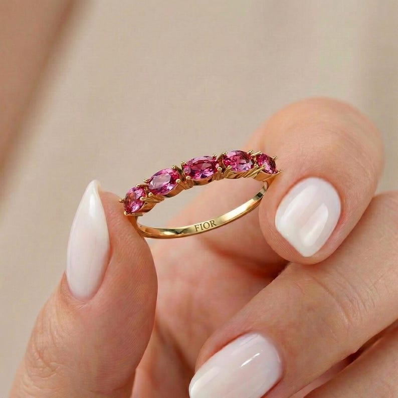 Op de afbeelding: Een gouden ring met een rij ovale roze edelstenen. De ring wordt vastgehouden in een hand met witte nagellak. Het woord "FIOR" is gegraveerd op de ringband. De ring is een sieraad.