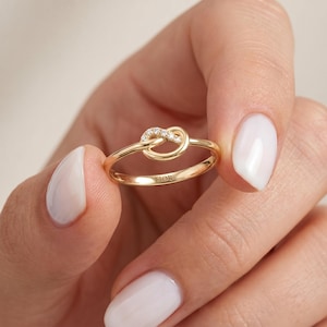 Könnte beinhalten: Ein goldener Ring mit einem zentralen Knotendesign, das mit kleinen, klaren, funkelnden Steinen verziert ist. Das Band ist glatt und poliert, wobei das Wort FIOR deutlich auf der Innenseite des Metalls eingraviert ist und eine feine Ästhetik zeigt.