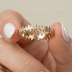 Puede incluir: Un anillo dorado con un diseño único. El anillo presenta una serie de barras verticales alrededor de la banda, creando un patrón geométrico moderno. La palabra "FIOR" está inscrita en el anillo. El anillo se sostiene entre dos dedos con esmalte de uñas blanco.