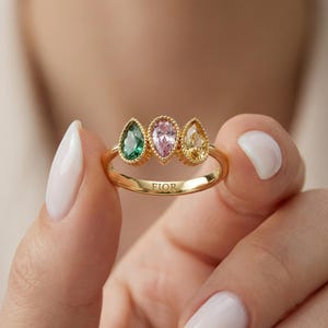 Puede incluir: Un anillo de oro con tres piedras preciosas en forma de lágrima en verde, rosa y amarillo. El anillo tiene una banda texturizada y la palabra "FIOR" está grabada en la banda.