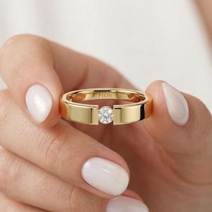 Könnte beinhalten: Ein goldener Ring mit einem einzelnen, runden, klaren Edelstein in der Mitte. Der Ring hat ein flaches Band und das Wort "FIOR" ist im Inneren eingraviert. Der Ring wird zwischen zwei Fingern gehalten.