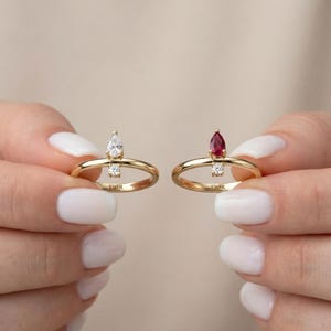 May include: Two gold rings, each with a distinctive gemstone. One ring has a pear-shaped white gemstone, the other a pear-shaped red gemstone. Both rings have a smaller, round gemstone below the larger stone. The rings are held up by a person.