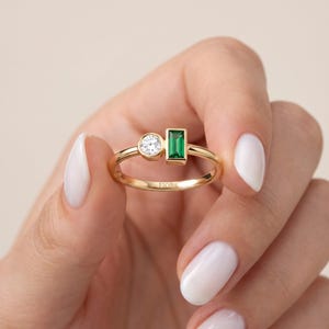 May include: A gold ring featuring a round brilliant-cut clear stone and a rectangular baguette-cut green stone set side-by-side on a thin polished gold band. The word FIOR is engraved on the inner surface of the gold band of the ring.
