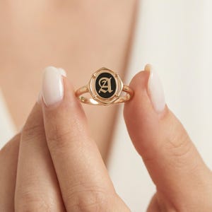 May include: A gold-colored ring with a black oval center featuring the letter "A" in a stylized font. The ring has a delicate band and a decorative setting around the black center. The ring is held up by a person's fingers.