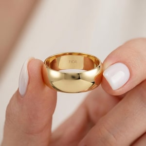 Könnte beinhalten: Ein polierter Goldring mit einer glatten, abgerundeten Außenfläche. Auf der Innenseite des Rings ist der Text FIOR in einer klaren, serifenlosen Schrift eingraviert. Das Metall hat eine hochglänzende, reflektierende Oberfläche.