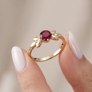 May include: A gold ring featuring a round, deep red gemstone set in the center, flanked by two leaf-shaped metal accents on each side. The inner band of the ring is engraved with the text FIOR in clear, capital letters, showcasing a polished finish.