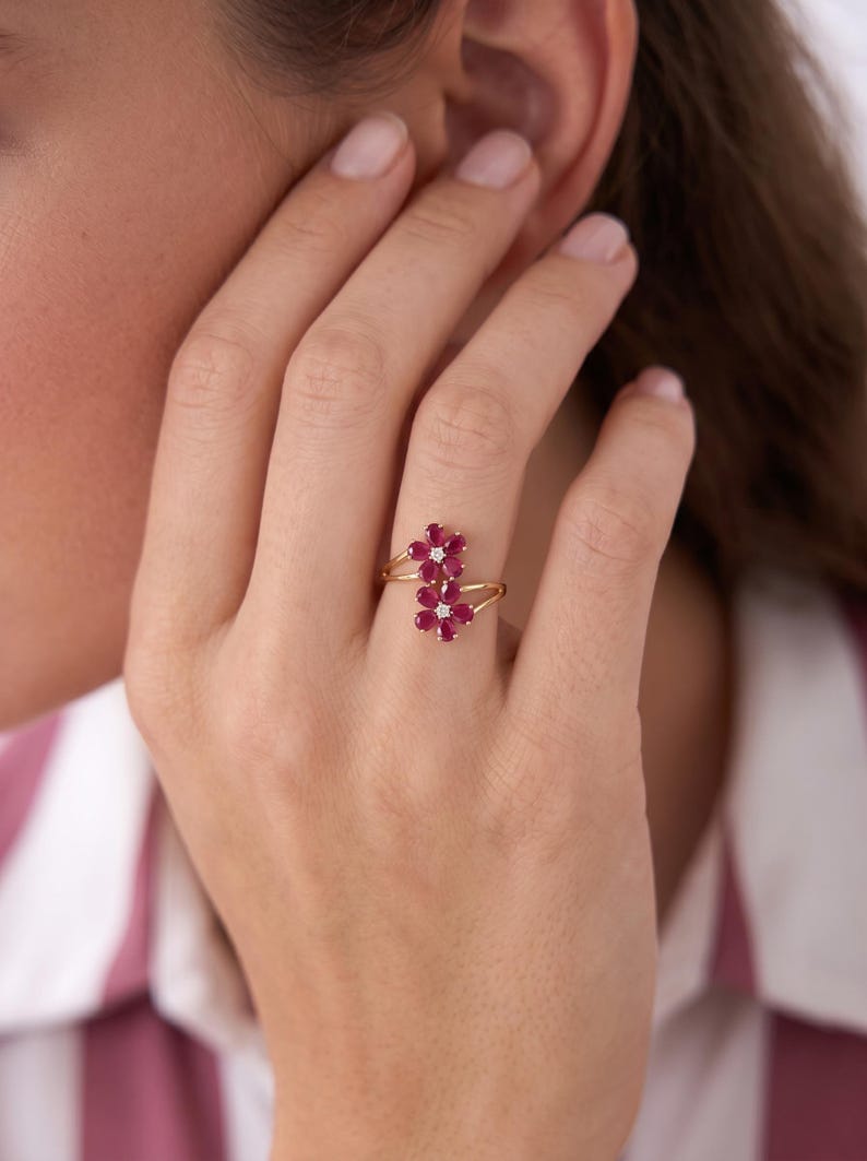 Puede incluir: Un anillo de oro con dos motivos en forma de flor, cada uno compuesto por cinco rub&iacute;es rojos de talla pera que rodean un peque&ntilde;o diamante blanco central. La banda es fina y dividida, conect&aacute;ndose a la base de cada dise&ntilde;o floral.
