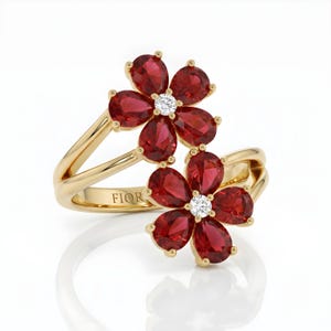 Puede incluir: Un anillo de oro con dos motivos en forma de flor, cada uno compuesto por cinco piedras rojas de talla pera que rodean una piedra blanca redonda central. La banda es de oro pulido e incluye el texto grabado FIOR en la parte interior.
