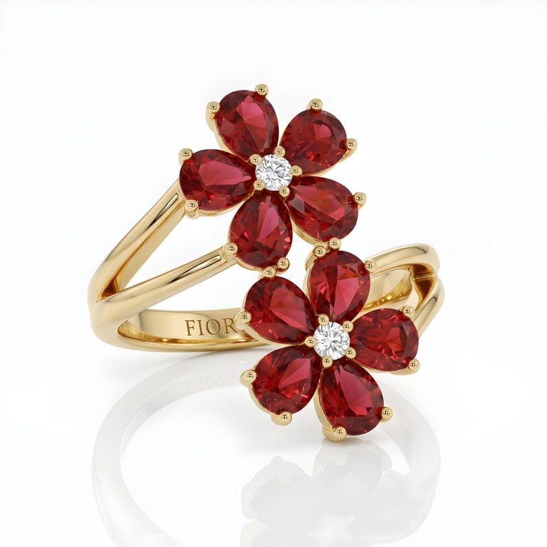 Puede incluir: Un anillo de oro con dos motivos en forma de flor, cada uno compuesto por cinco piedras rojas de talla pera que rodean una piedra blanca redonda central. La banda es de oro pulido e incluye el texto grabado FIOR en la parte interior.