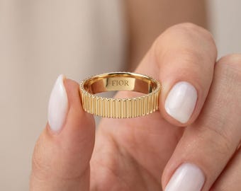 Puede incluir: Un anillo de tono dorado que presenta una textura acanalada vertical a lo largo de la banda exterior. El interior del anillo es liso y está claramente grabado con el texto FIOR. La banda tiene un ancho constante y un acabado pulido.
