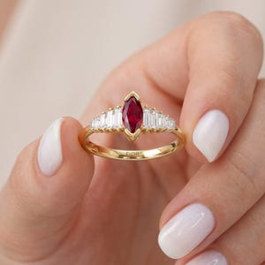 May include: A gold ring featuring a central marquise-cut red gemstone flanked by three baguette-cut clear stones on each side. The band is polished gold and includes the engraved text FIOR on the inner surface of the ring's metal band.