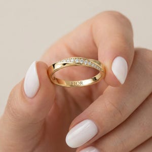 May include: A gold-colored ring with a twisted design, set with small, clear gemstones. The ring is held up by a person with white nail polish. The word "FIOR" is engraved on the inside of the ring.