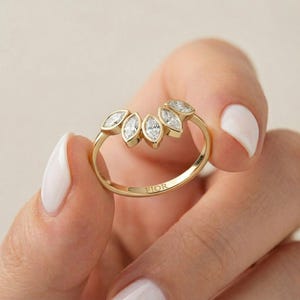 Könnte beinhalten: Ein goldener Ring mit einem Blattdesign, besetzt mit sechs Diamanten im Marquiseschliff. Der Ring wird in einer Hand mit weißem Nagellack gehalten. Das Wort "FIOR" ist auf dem Ringband eingraviert.