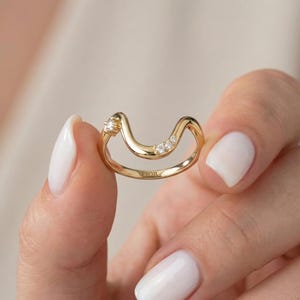 May include: A gold ring with a distinctive wave shape, set with three small, clear gemstones. The ring is held by a person with white nail polish. The word "FIOR" is engraved on the ring.