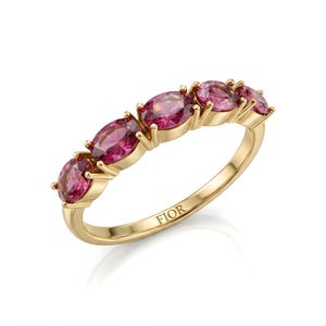 Op de afbeelding: Een gouden ring met vijf ovale roze edelstenen. De edelstenen zijn in een rij geplaatst, met een gouden band. De ring staat tegen een witte achtergrond. Het woord "FIOR" is op de band gegraveerd.