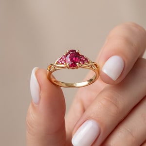 May include: A gold ring featuring a central oval-cut deep red gemstone flanked by three smaller red stones on each side. The band has a delicate, swirling metal design and is engraved with the word FIOR on the inner surface of the ring.