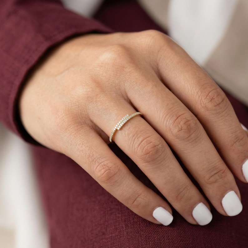 May include: A gold ring featuring a double band design, each adorned with small, clear gemstones. The ring is displayed on a finger, with a maroon sleeve and white nails visible.