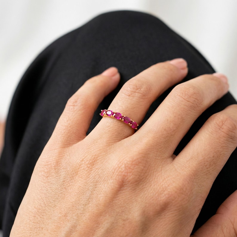 Op de afbeelding: Een gouden ring bezet met ovaal geslepen, levendige rode edelstenen. De ring wordt op een hand gedragen, waarbij de kleur van de edelstenen en het ontwerp van de ring worden getoond. De band van de ring is dun en gepolijst goud.