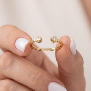 Peut inclure: Une bague ouverte en or ornée de deux petites pierres rondes et transparentes serties clos à chaque extrémité de l'anneau. L'anneau est en or poli et porte l'inscription FIOR gravée en lettres capitales sur la surface extérieure de la tige.