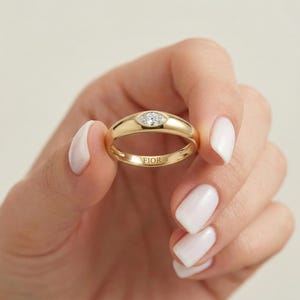 May include: A gold ring featuring a marquise-cut diamond. The ring has a polished band and the word "FIOR" is engraved inside. The ring is held up by a person with white nail polish.