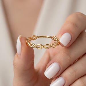 Peut inclure: Une bague dorée avec un motif entrelacé est tenue entre des doigts avec du vernis à ongles blanc. La bague porte l'inscription "FIOR". Le motif de la bague est un motif continu de boucles entrelacées.
