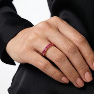 Op de afbeelding: Een gouden ring met een reeks ovaal geslepen, helderrode edelstenen. De ring is gezet in een gouden band en wordt getoond op een vinger. De edelstenen zijn gelijkmatig verdeeld, waardoor een doorlopende lijn van kleur ontstaat.