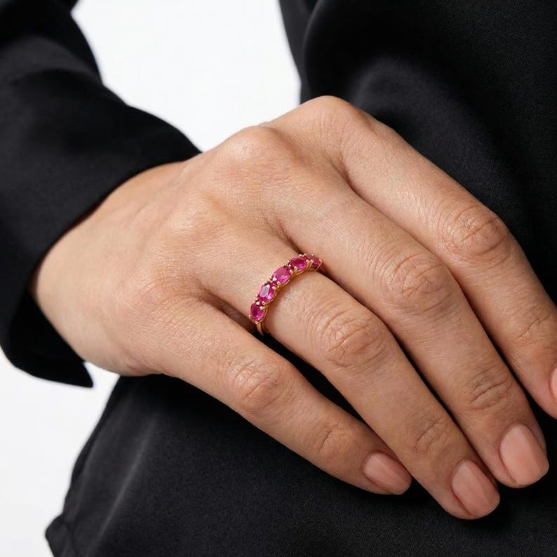 Op de afbeelding: Een gouden ring met een reeks ovaal geslepen, helderrode edelstenen. De ring is gezet in een gouden band en wordt getoond op een vinger. De edelstenen zijn gelijkmatig verdeeld, waardoor een doorlopende lijn van kleur ontstaat.