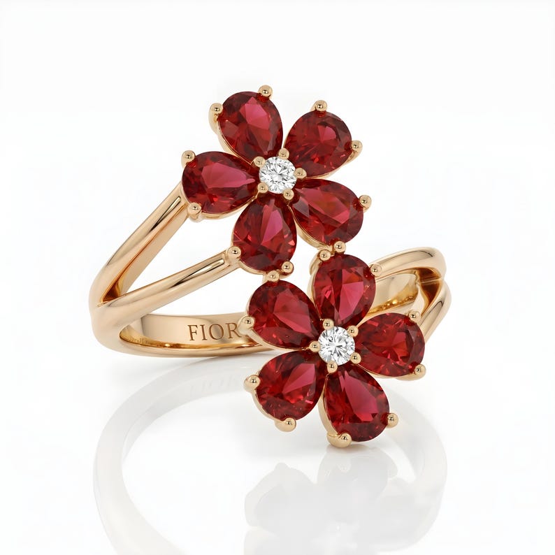 Puede incluir: Un anillo de oro con dos motivos en forma de flor, cada uno compuesto por cinco piedras rojas en talla pera que rodean un diamante blanco redondo central. La banda est&aacute; dividida y pulida, con la palabra FIOR grabada en el interior del metal.