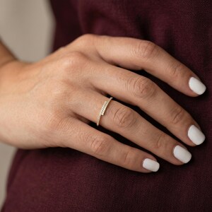 May include: A gold ring with an unusual, asymmetrical design. The ring has a band of small, sparkling stones set along a diagonal line. The ring is shown on a hand with white nail polish, against a dark red background.