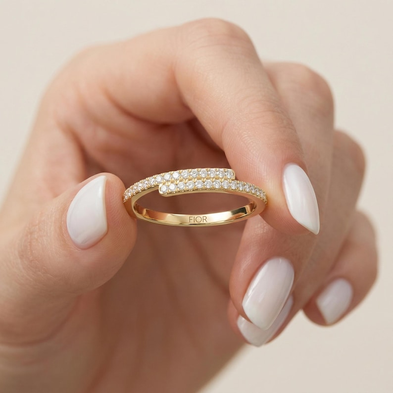May include: A gold ring with a distinctive design, set with small, sparkling diamonds. The ring has a curved, overlapping design, with the word "FIOR" engraved on the band. The ring is held up by a hand with white nail polish.