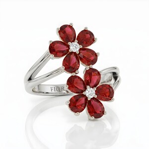Puede incluir: Un anillo de plata con dos motivos en forma de flor, cada uno compuesto por cinco piedras rojas de talla pera que rodean una piedra blanca redonda central. La banda es dividida y pulida, con el texto FIOR grabado en la superficie interior.