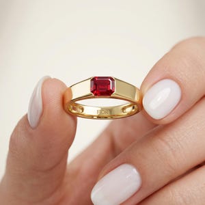 Puede incluir: Un anillo de oro con una piedra preciosa roja de corte esmeralda rectangular en el centro. La banda es lisa y pulida, con la palabra FIOR grabada en la superficie interior de la banda de metal, mostrando un diseño limpio y sencillo.