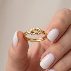 Può includere: Un anello color oro con un design a nodo. L'anello presenta una doppia fascia e un elemento centrale a forma di nodo. L'anello è tenuto in una mano con smalto bianco per unghie. È un gioiello.
