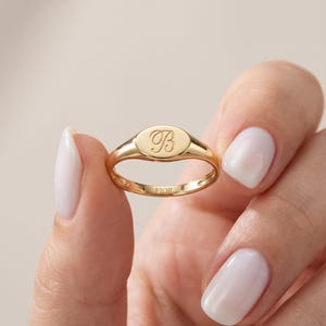 May include: A polished gold signet ring featuring an oval face engraved with a cursive letter B. The interior of the band is clearly stamped with the text FIOR. The ring has a smooth, reflective metallic finish and a slender, rounded band design.