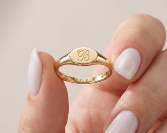 May include: A polished gold signet ring featuring an oval face engraved with a cursive letter B. The interior of the band is clearly stamped with the text FIOR. The ring has a smooth, reflective metallic finish and a slender, rounded band design.