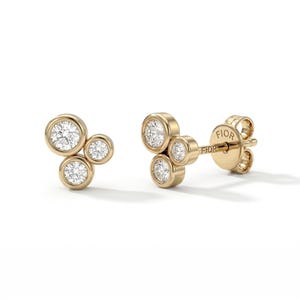 May include: A pair of gold stud earrings, each with three round, bezel-set diamonds. The earrings have a cluster design, with the diamonds arranged in a curved pattern. The post of one earring is stamped with the word "FIOR".