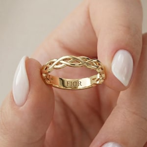 May include: A gold ring featuring a braided, woven-style band design on the top half. The interior surface of the smooth gold band is clearly engraved with the text FIOR in capital letters, visible within the centre of the ring's circular opening.