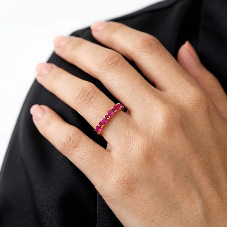 Op de afbeelding: Een gouden ring met een rij ovaal geslepen, helderrode edelstenen. De ring zit om een vinger, met de hand tegen een zwart kledingstuk. De edelstenen zijn gelijkmatig verdeeld en glinsteren.
