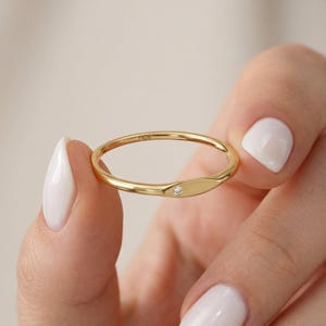 May include: A gold ring featuring a small diamond set in a smooth, oval design. The ring is held between two fingers, highlighting its polished surface and delicate details. The word "FICR" is engraved on the ring.