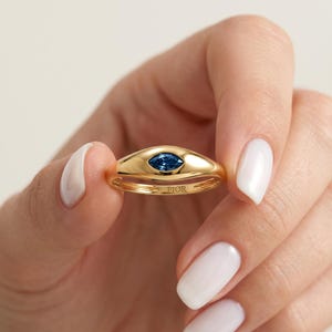 May include: A gold ring featuring a blue gemstone in an oval setting. The ring has a smooth, polished finish, with the word "FIOR" engraved on the band. The ring is held by a person with white nail polish.