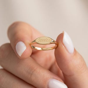 May include: A gold oval signet ring, engraved with the name "Jessica". The ring is held up, showing the engraved name. The ring is a classic design, suitable for everyday wear.