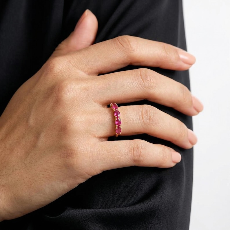Op de afbeelding: Een gouden ring met een rij ovaal geslepen, levendige rode edelstenen. De ring wordt getoond op een vinger, waarbij het gepolijste metaal en de rijke kleur van de stenen worden benadrukt. De ring is een sieraad.